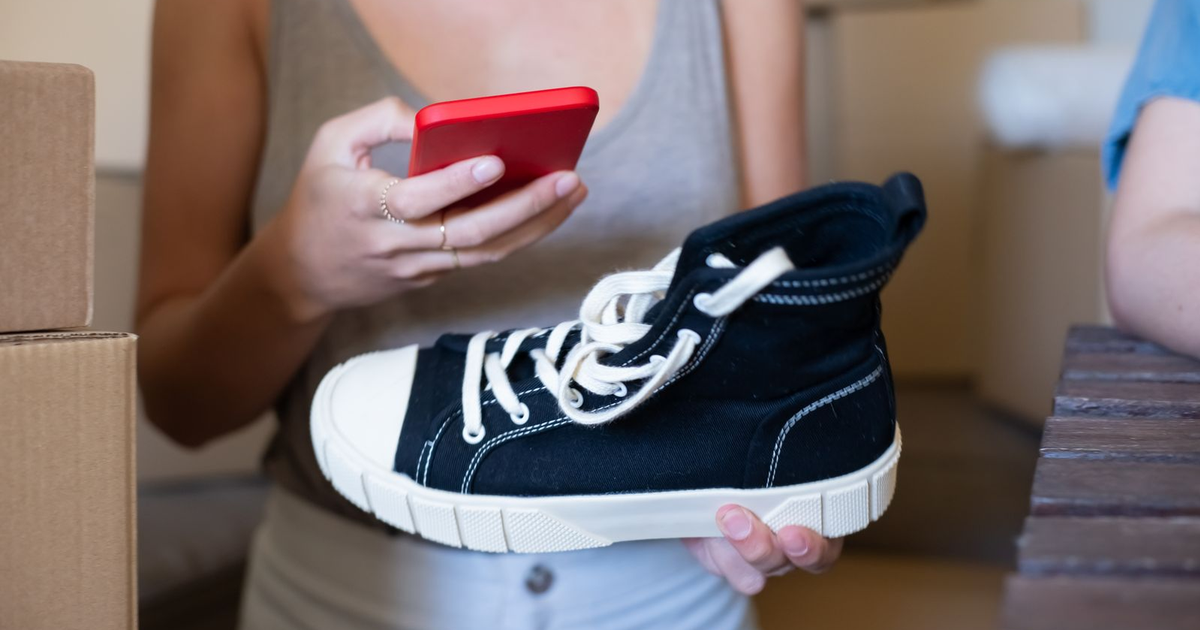 vendedora online con una zapatilla y un móvil entre cajas de envío: un producto que los compradores pueden ver ahora en una ficha dentro de la app